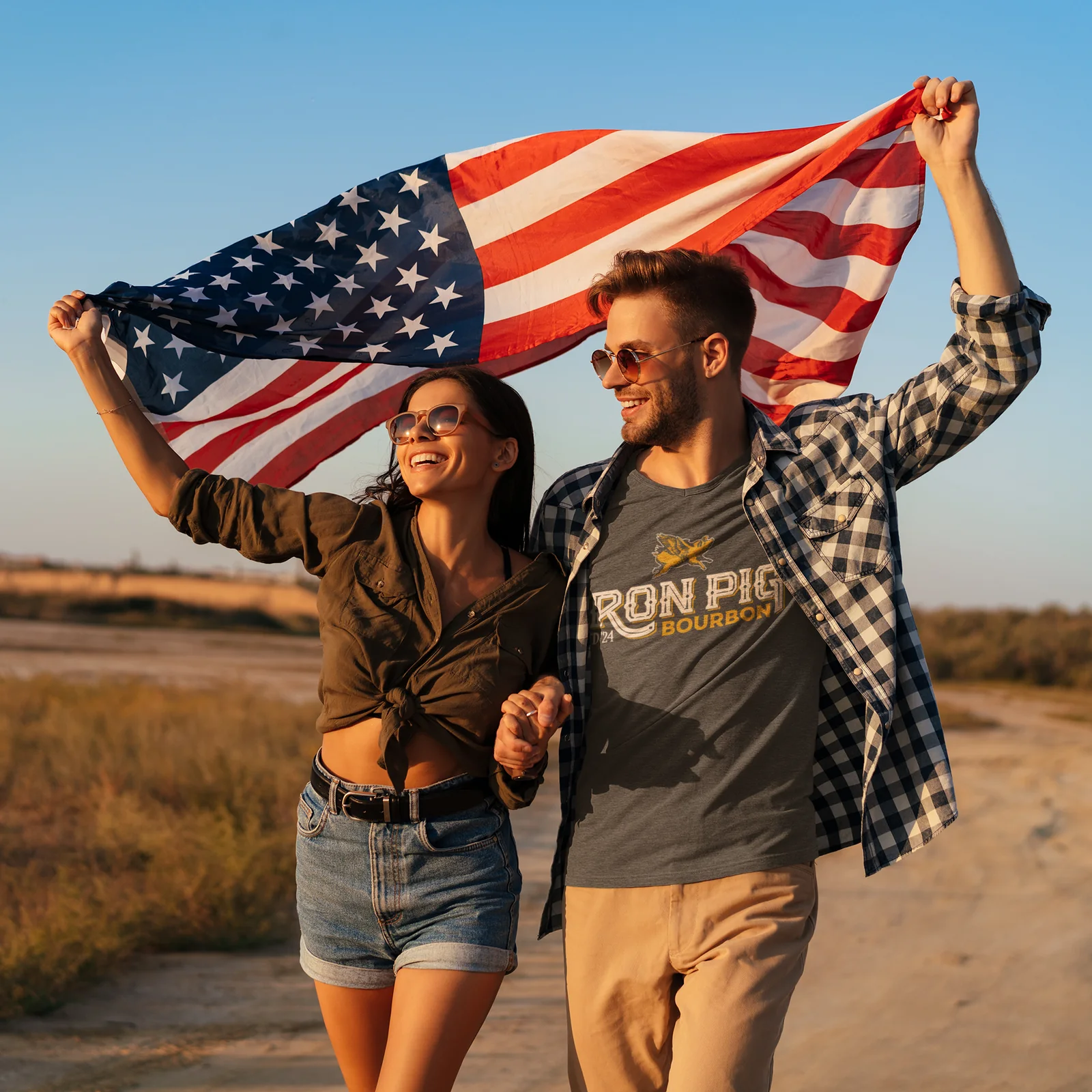 Couple walking a dirt road holding an American flag overhead, the man in a gray Iron Pig Bourbon T-shirt